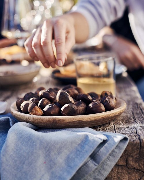 Hand greift geröstete Maronen von Holzschale auf rustikalem Tisch
