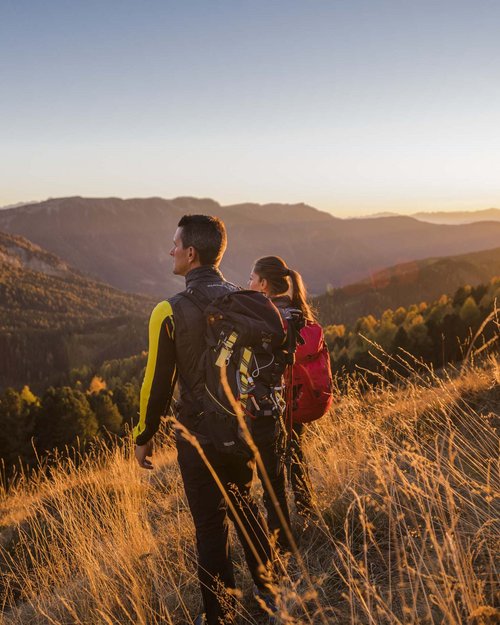 Two hikers with backpacks watch the mountain landscape at sunset.