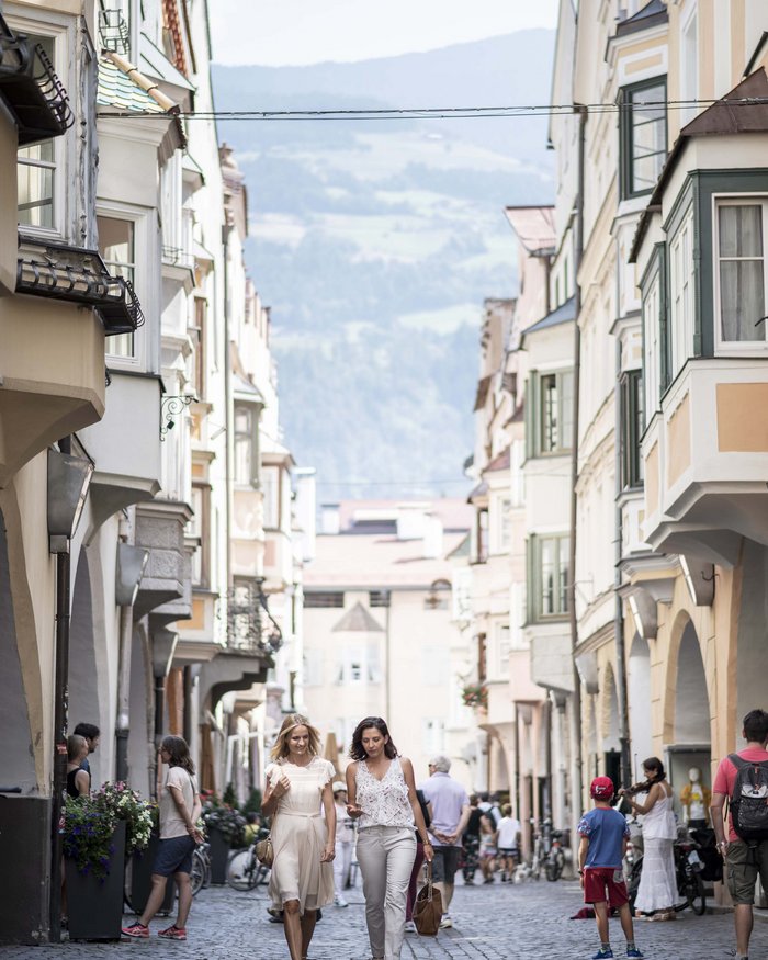 Zwei Frauen gehen durch eine belebte Gasse mit historischen Gebäuden und Bergen im Hintergrund