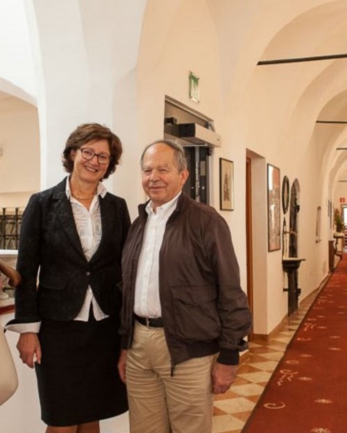 Two people standing in a hallway with vaulted ceiling and red carpet.