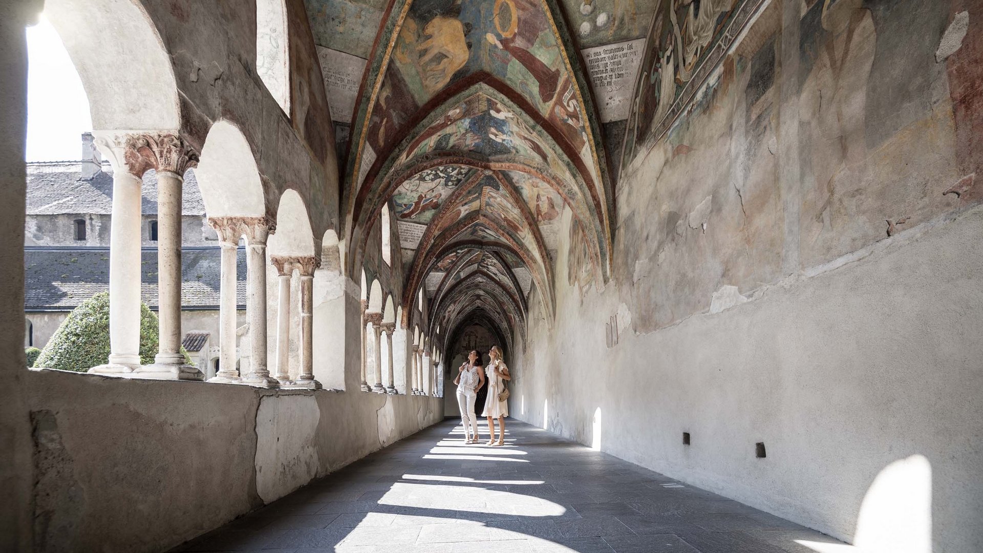 Two women observing frescoes in a vaulted cloister with columns and arches
