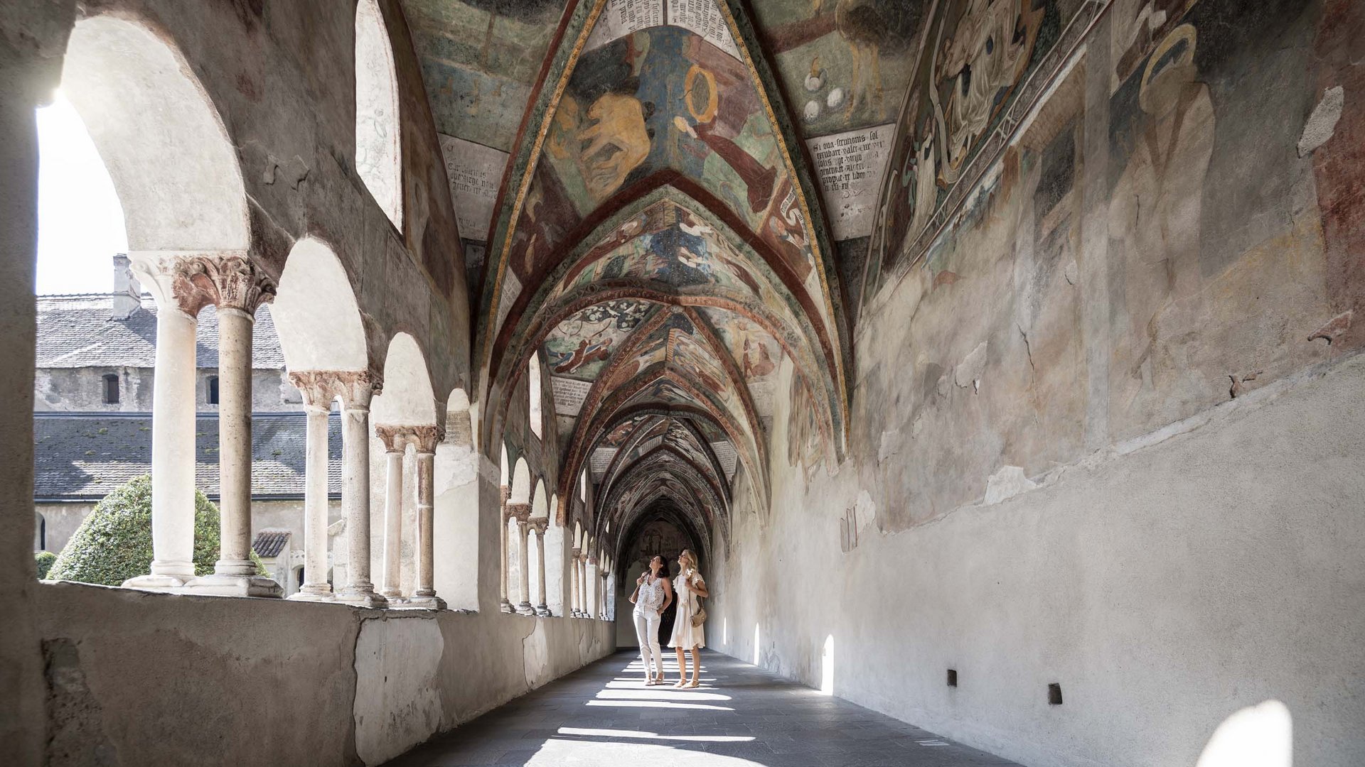 Two women observing frescoes in a vaulted cloister with columns and arches