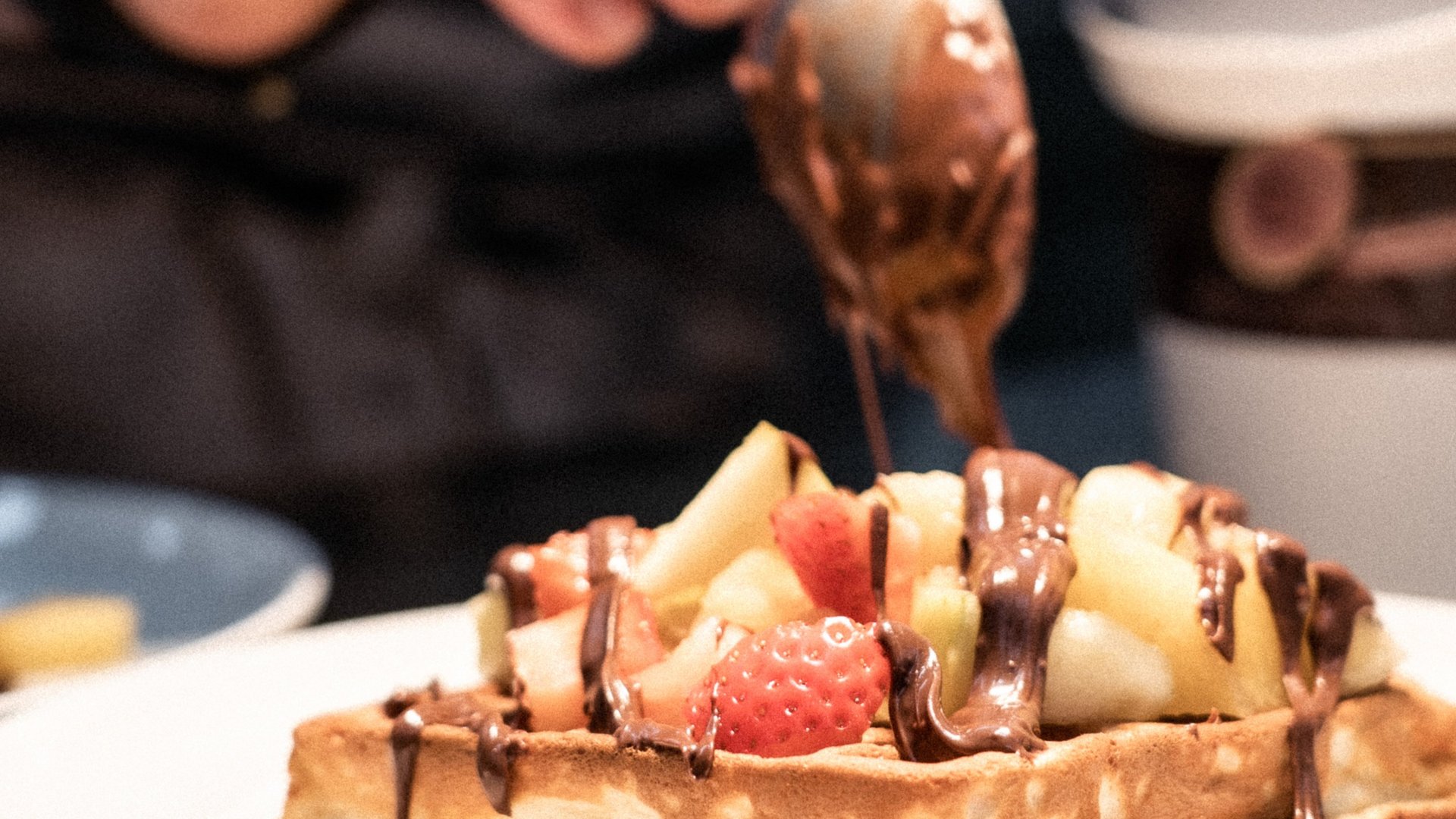 Hand drizzling chocolate sauce on waffle topped with fruit on white plate