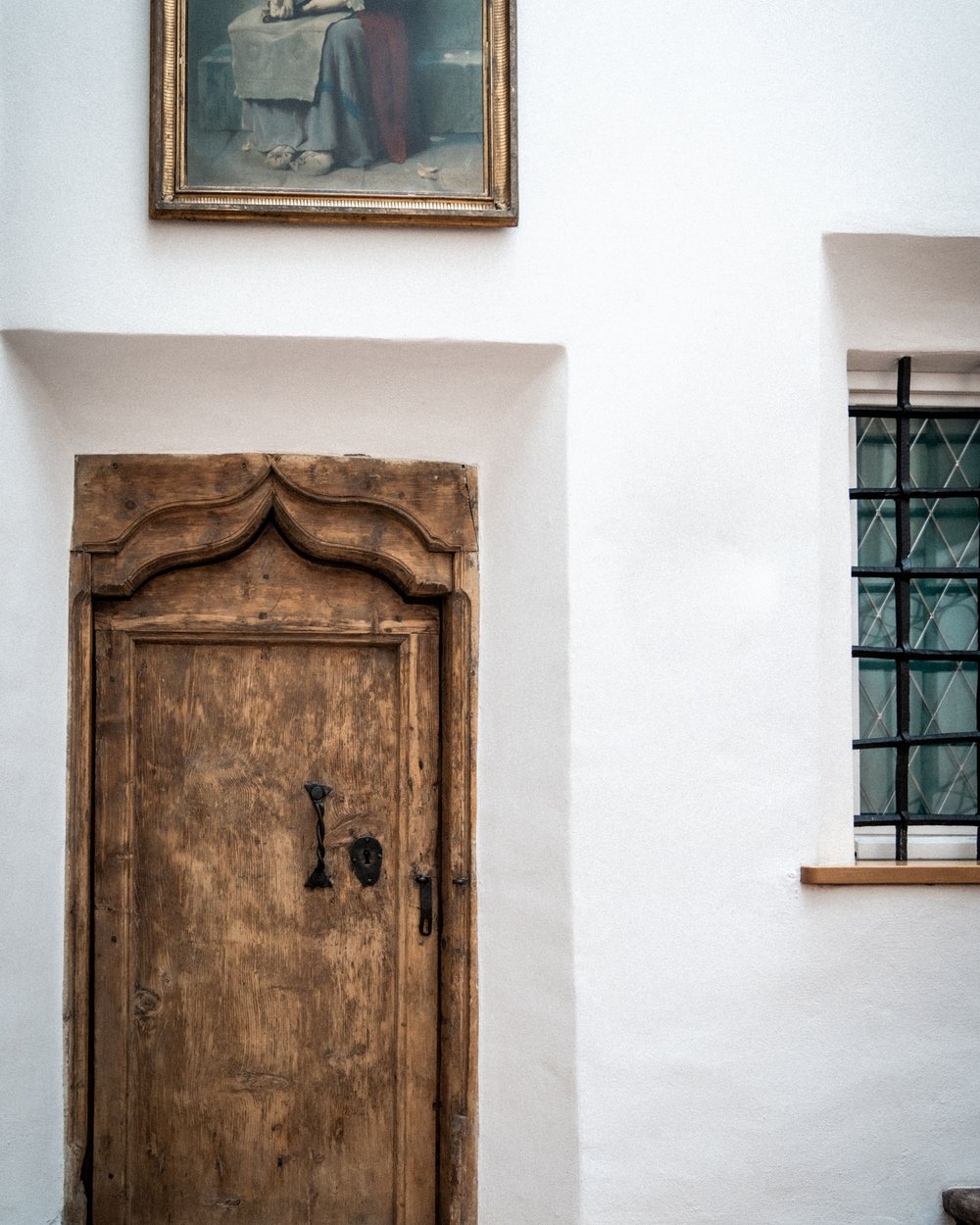 Old ornate wooden door in white wall with painting above and window beside it