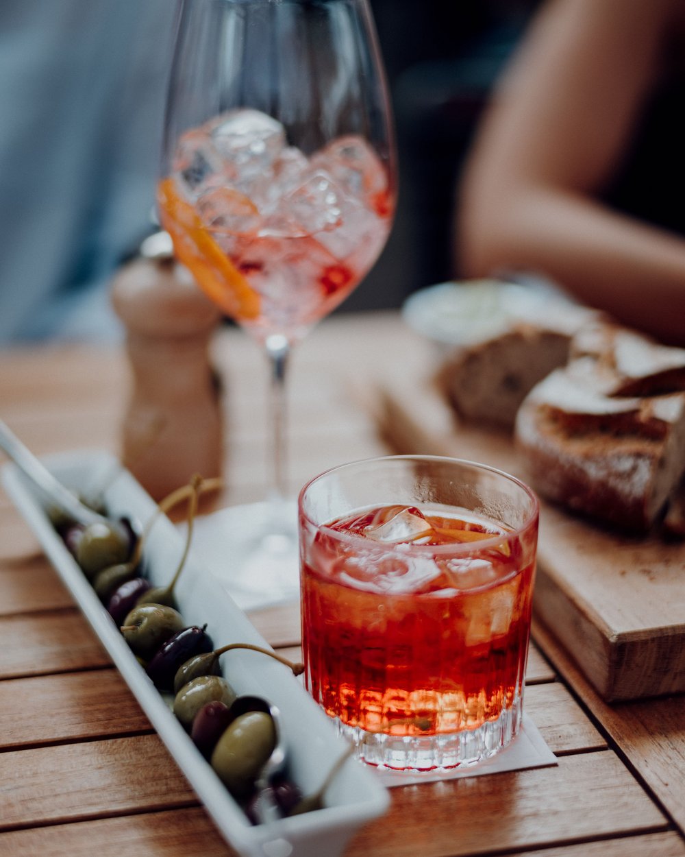 A glass of red drink, another glass with ice, and a dish of olives on a wooden table