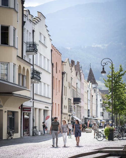 Menschen spazieren in einer Alpenstadt mit bunten Häusern und Bergen im Hintergrund