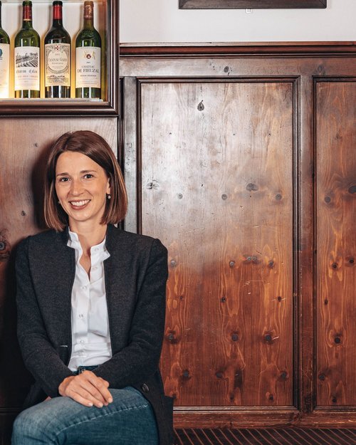 Two smiling people sitting in front of wood paneling with wine bottles above