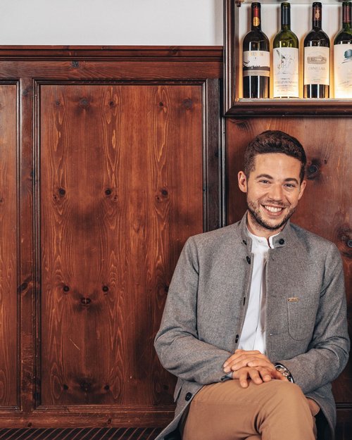 Two smiling people sitting in front of wood paneling with wine bottles above