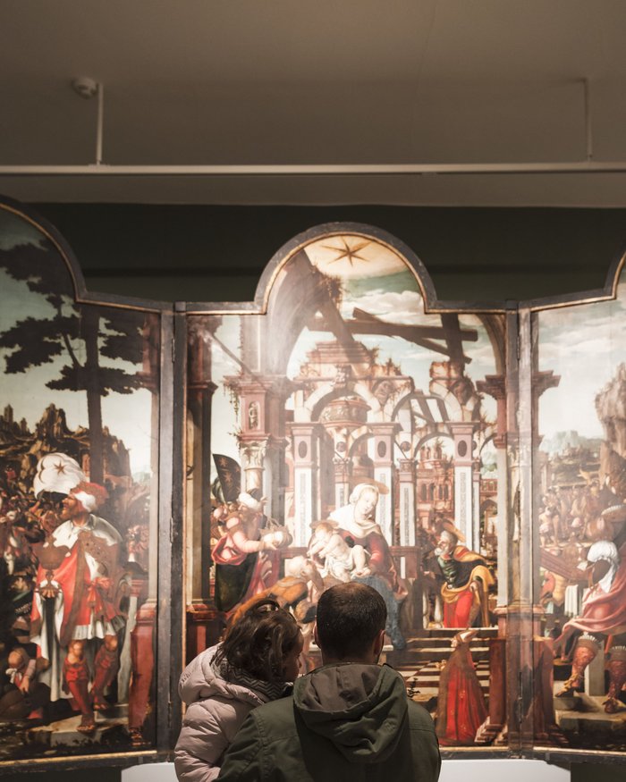 A man carries a child in front of a Renaissance painting in an art gallery