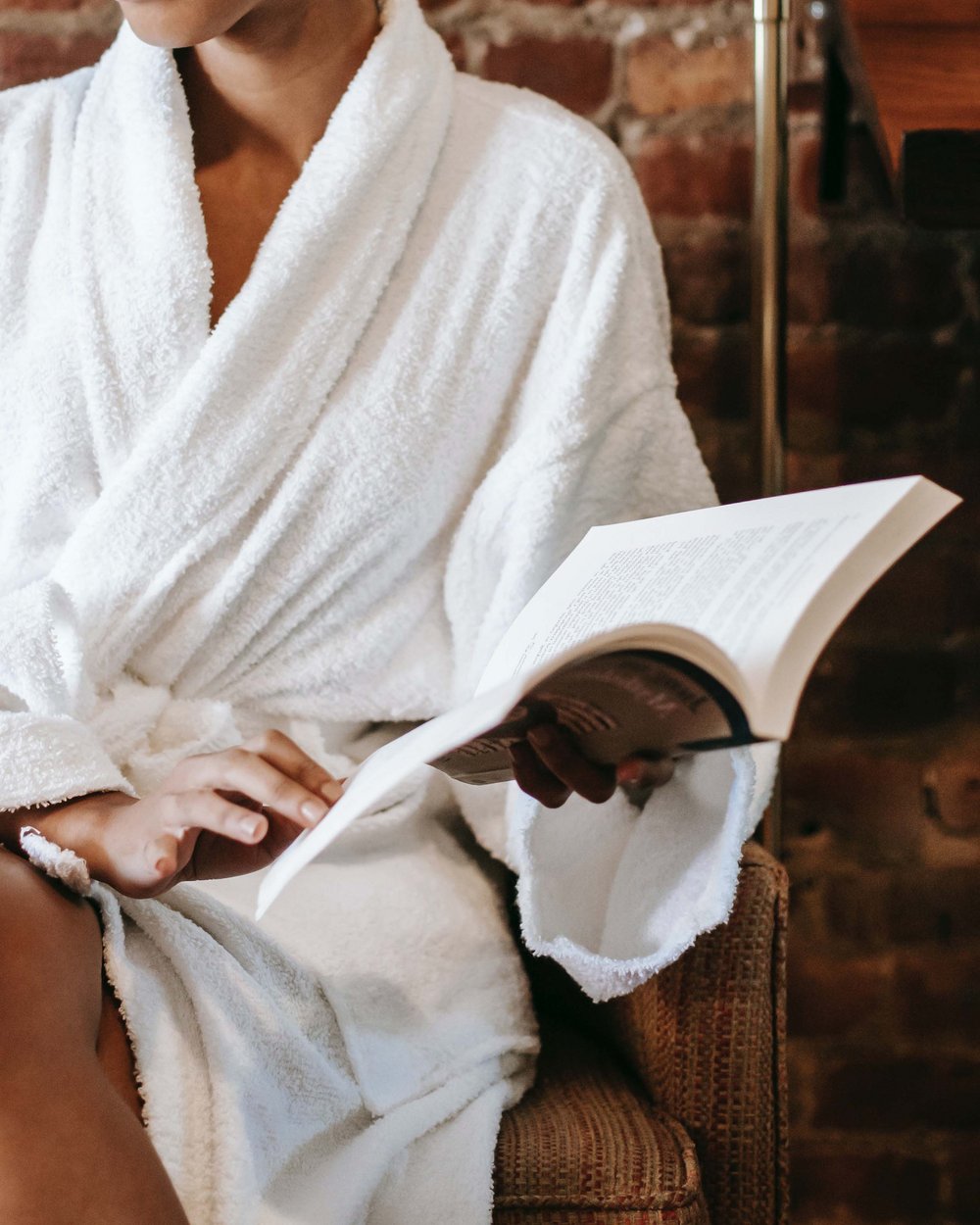 Frau im weißen Bademantel liest Buch auf Stuhl vor Backsteinwand